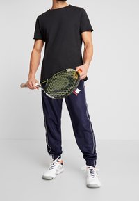 Czarna bawełniana koszulka, luźny krój; granatowe spodnie dresowe z białymi paskami po bokach; trzyma rakietę tenisową; białe buty sportowe z pomarańczowymi akcentami.
