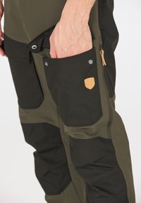 Cargo-housut ovat oliivinvihreät ja mustat, ja niissä on useita taskuja, nepparikohteita ja nahkakoristeita. Kankaan tekstuuri näkyy.