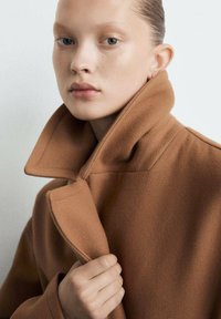 Bruine wollen jas met een hoge kraag, gestructureerde stof en een opvallende plooi. Een hand grijpt de kraag vast en omlijst het gezicht. Neutrale achtergrond.