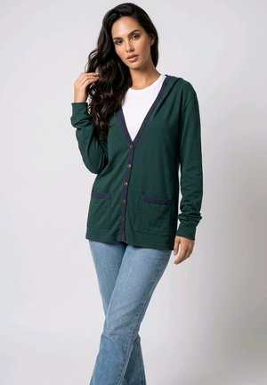 Mujer con cabello largo y oscuro que lleva un cárdigan verde con botones sobre una camisa blanca y vaqueros azul claro, de pie frente a un fondo sencillo.