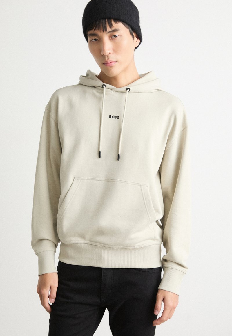 Sudadera beige con bolsillo canguro, capucha con cordón y logo negro "BOSS". Lleva puesta sobre pantalones negros, con puños y dobladillo acanalados. Textura suave.