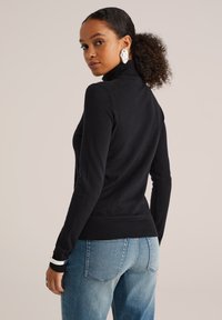 Femme aux cheveux bouclés en queue de cheval portant un pull col roulé noir, un jean bleu clair, de grandes boucles d'oreilles en argent et un bracelet blanc.