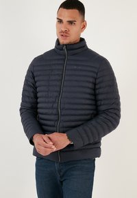 Giacca puffer blu navy con colletto alto, design trapuntato orizzontale, zip frontale e tasche laterali. Texture liscia e aspetto leggero.