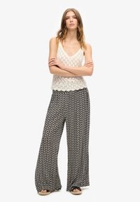 Top sin mangas de punto ligero con un patrón de encaje en color crema, combinado con pantalones amplios negros que presentan un patrón ovalado blanco repetitivo. Sandalias planas visibles.