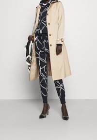 Beige trenchcoat, marinblå hoodies med vit kedjemönster, matchande sweatpants och svarta lackskor med geometriska detaljer.