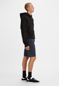 Hanorac negru cu buzunar tip cangur, purtat cu pantaloni scurți bleumarin. Adidași negri cu tălpi albe și șosete până la gleznă completează ținuta. Vedere laterală.