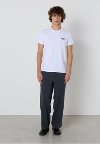 Mladenič stoji obrnjen naprej, oblečen v belo majico z majhnim logotipom, ohlapne temno sive hlače in črne škornje proti enostavni ozadju.