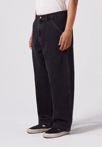 Jean en denim noir avec une coupe décontractée, design à cinq poches, surpiqûres visibles et fermeture par bouton à la taille. Porté avec des baskets.