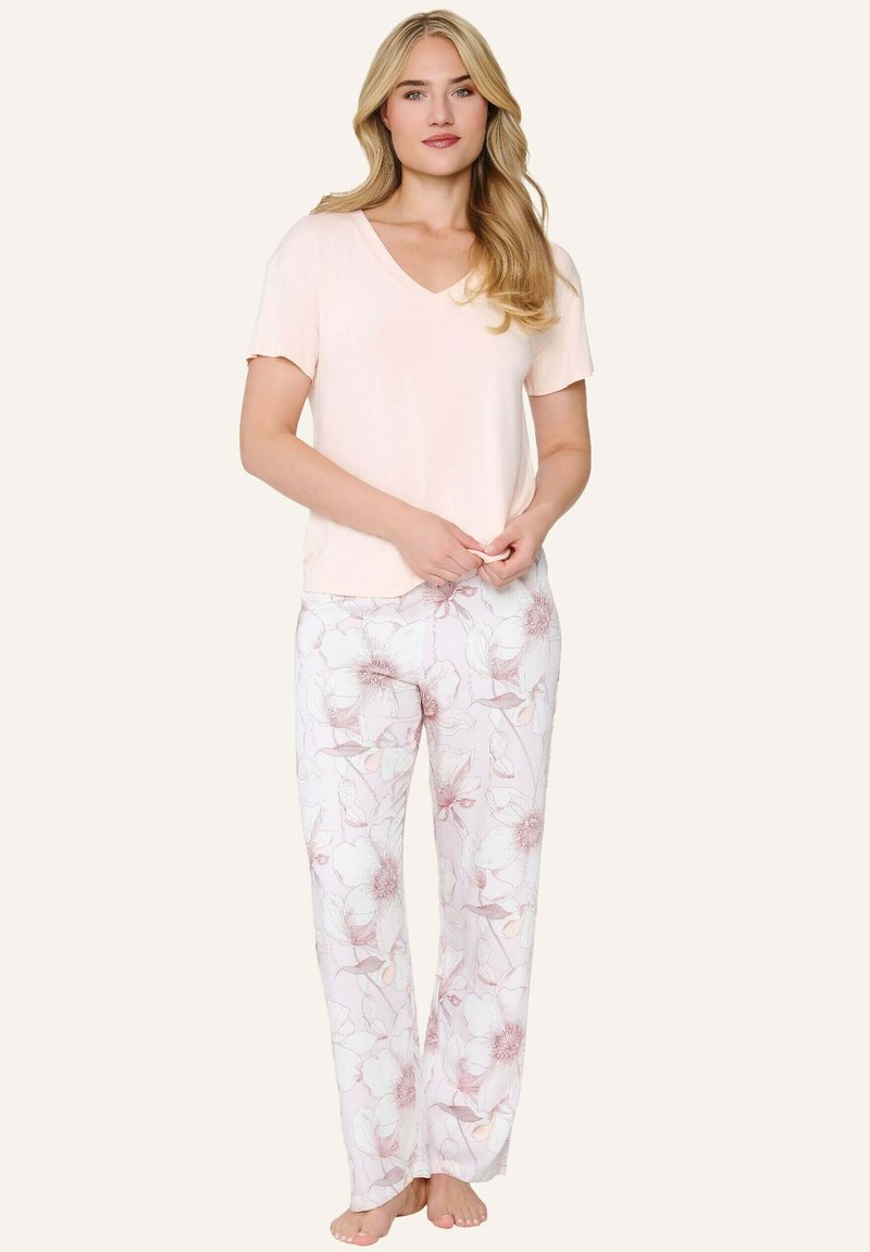 Femme aux longs cheveux blonds portant un haut rose clair à manches courtes et un pantalon blanc à motif floral, debout pieds nus sur un fond uni.