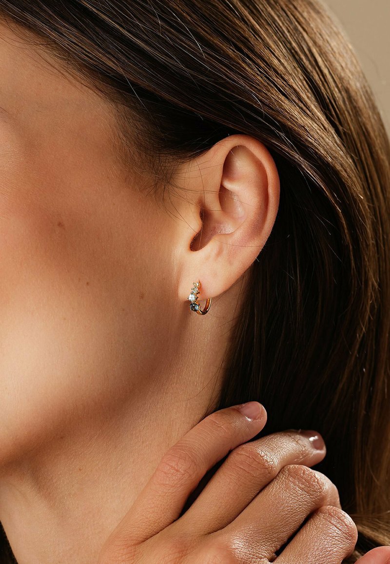 Guldringar med en ädelsten, prydda med en liten blå sten och klara rhinestones, visade på en modells öra.