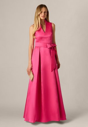 Femme porte une robe en satin rose vif sans manches avec un décolleté en V, une large ceinture nouée en nœud, tenant une pochette beige sur un fond uni.