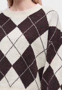 Femme portant un pull en tricot à motif losanges crème et marron foncé avec un col rond et des manches longues.