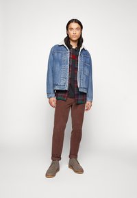Jeansjack met een witte sherpa-kraag, gelaagd over een groen en rood geruit shirt, gecombineerd met bruine corduroybroek en grijze laarzen.