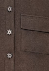 Close-up rjave volnene jakne, ki prikazuje tri sivo gumbe, nameščene navpično, ter veliko pravokotno žep s pokrovom.