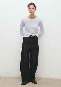 Weite schwarze Denim-Hosen mit gelben Nähten, kombiniert mit einem hellgrauen Langarmshirt. Das Modell steht vor einer weißen strukturierten Wand.