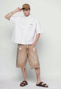 Giovane uomo che indossa una maglietta bianca a maniche corte, larga, pantaloni lunghi beige strappati, un cappellino marrone e sandali marroni, in piedi con una mano che tocca il cappello.
