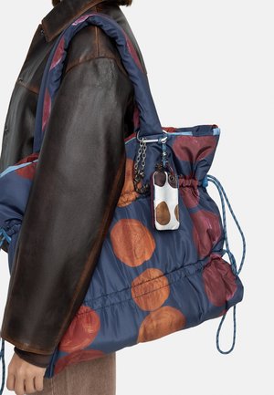 Osoba nosząca ciemnobrązową skórzaną kurtkę, niosąca dużą niebieską torbę typu tote z pomarańczowo-maronowymi, okrągłymi wzorami oraz wiszącą breloczek.