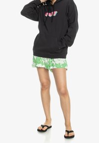 Sweat à capuche noir avec un logo graphique coloré, associé à une jupe blanche ornée de motifs de feuilles vertes et des tongs noires.