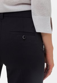 Černé kalhoty na míru vyrobené z tkaniny s hladkou texturou. Mají zadní kapsu s knoflíkovým zapínáním a diskrétním logem.