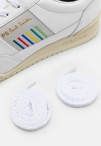 Zapatilla blanca con detalles de rayas multicolores y un logo, acompañada de dos cordones blancos bien enrollados sobre una superficie blanca.