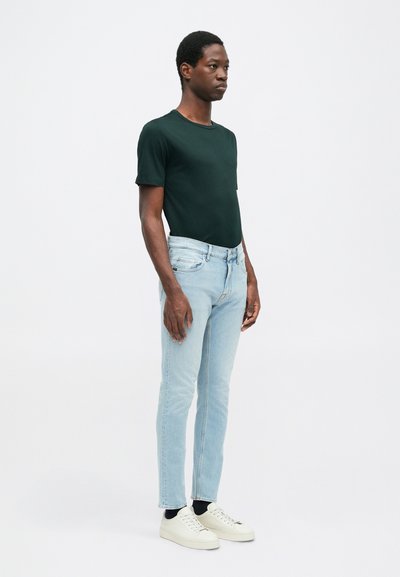 T-shirt s krátkym rukávom tmavozelenej farby, svetlomodré priliehavé džínsy a biele tenisky. Model stojí proti jednoduchému svetlému pozadiu.