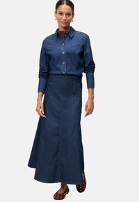 Koszula dżinsowa z ostrym kołnierzykiem, dopasowanym fasonem i zapięciem na guziki. Długa spódnica dżinsowa z bocznymi rozcięciami i rozszerzanym dołem. Brązowe buty z ćwiekami.