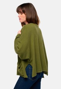 Pull oversized vert olive avec épaules tombantes, texture côtelée et fentes sur les côtés, associé à un jean bleu foncé. Vue de côté affichée.