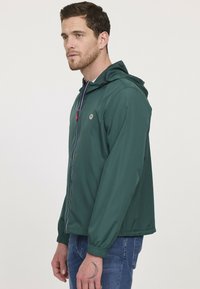 Grüne leichte Jacke mit Kapuze, Frontreißverschluss, elastischen Bündchen und Logo auf der Brust. Glatte Textur mit kontrastierenden Reißverschlüssen.