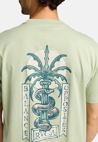 Camiseta de algodón verde claro con un gráfico central de una serpiente entrelazada y una palma, con las palabras "Equilibrio" y "Opuestos."