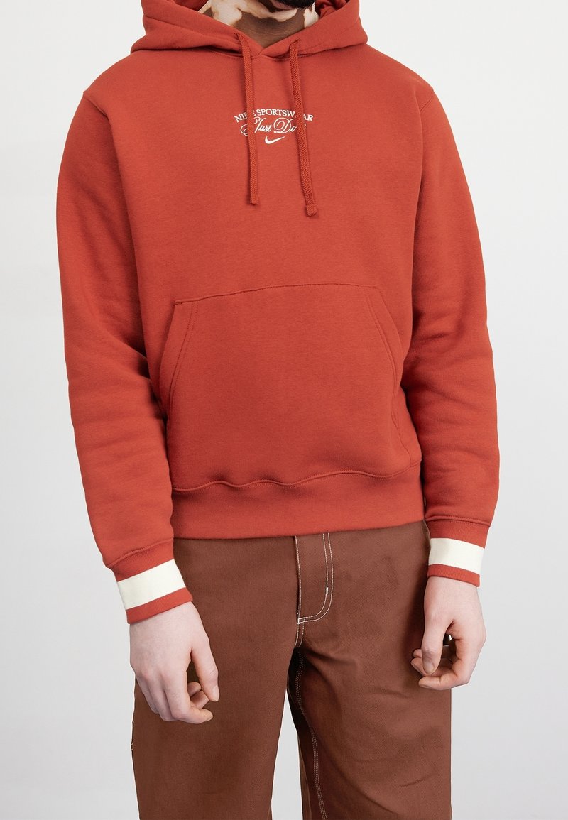 Röd huvtröja med en känguruficka, prydd med vit broderad text och kontrasterande randiga muddar. Kombinerad med bruna byxor.