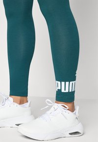 Vihreät legginsit, joissa on sileä pinta ja valkoinen "PUMA" -painatus nilkassa, yhdistettynä valkoisiin urheilukenkiin.