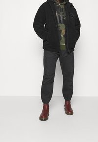 Svart, fluffig zip-up jacka över en grön kamouflage-hoodie, kombinerad med lösa svarta byxor och vinröda ankelstövlar.