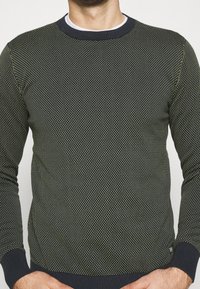 Tummanvihreä neulepaita, jossa on pieniä keltaisia pisteitä muodostava tekstuuri, resorinapit ja -helma sekä pyöreä pääntie. Sopii rentoon käyttöön.