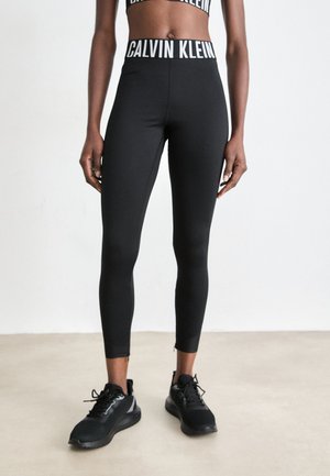 Persona che indossa leggings neri Calvin Klein e un reggiseno sportivo abbinato, in piedi su un pavimento chiaro, scarpe da ginnastica nere visibili.