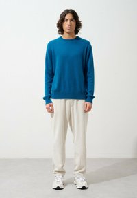 Pull en tricot turquoise avec un col rond, associé à un pantalon texturé crème. Le mannequin se tient devant un fond clair.