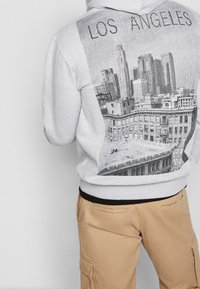 Felpa grigia con un grafico nero di uno skyline cittadino e la scritta "LOS ANGELES". Polsini e orlo a costine con una vestibilità rilassata. I pantaloni sono beige.