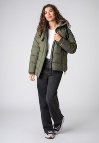 Gewatteerd jack in olijfgroen met een doorgestikt ontwerp, rits aan de voorkant en verstelbare capuchon. Gecombineerd met zwarte jeans met wijd uitlopende pijpen en sneakers.