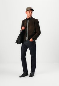 Brown suede jacket with a zip closure, grey shirt, dark denim jeans, and black leather boots. Black shoulder bag and plaid cap.