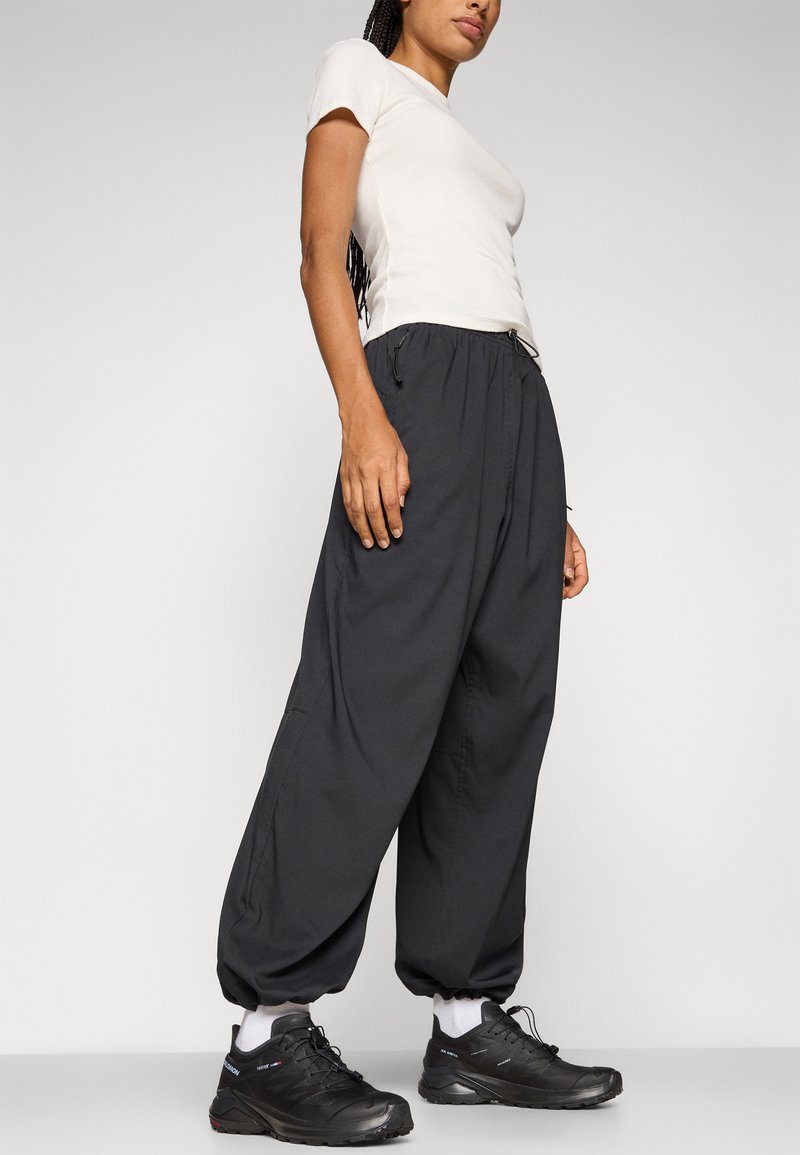 Schwarze, lockere Hosen mit elastischen Bündchen und einem Kordelzugbund, kombiniert mit einem eng anliegenden, hellen Oberteil und schwarzen Sportschuhen.