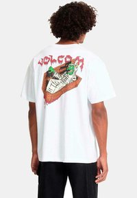 Persona con una camiseta blanca que tiene un gráfico colorido de un monstruo verde envuelto en vendas dentro de un ataúd, con el texto rojo "VOLCOM" arriba.