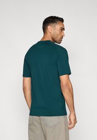 Türkizkék pamut póló rövid ujjakkal, kerek nyakkivágással és finom vállvarrás részletekkel. Világos bézs színű farmernadrággal viselve.