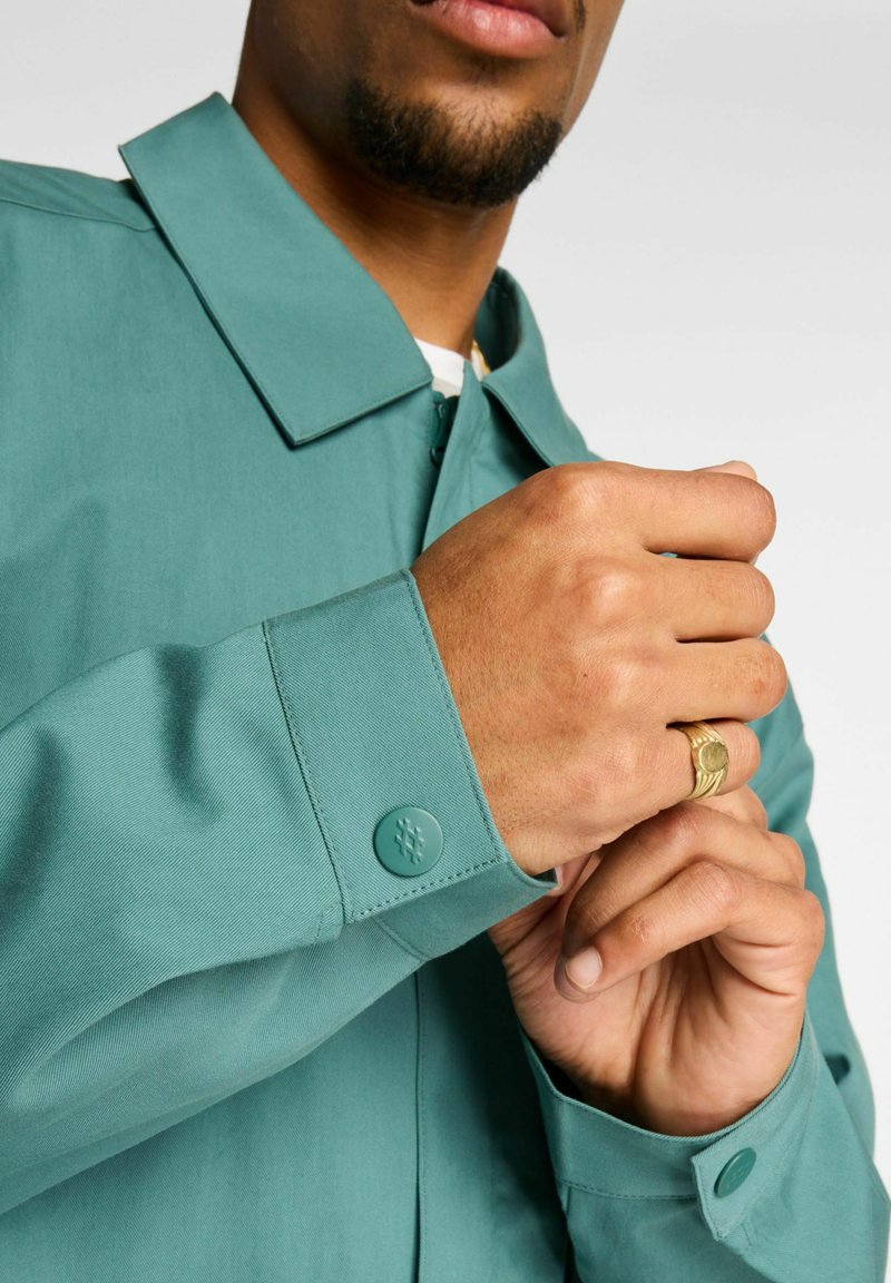 Hellgrüne Jacke mit Kragen, geknöpften Manschetten und einer glatten Textur. Verfügt über ein auffälliges Knopfdetail am Ärmel.