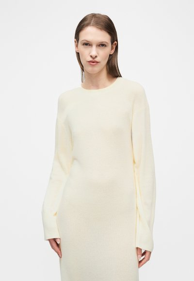 Γυναίκα με ίσια καστανά μαλλιά φοράει μακρύ κρεμ πλεκτό φόρεμα με μακριά μανίκια, στέκεται μπροστά σε απλό λευκό φόντο.