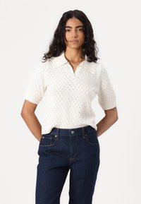 Knitted white pullover with a diamond pattern, short sleeves, and a flat collar. Paired with dark blue denim pants.