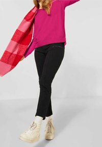 Fuchsijový svetr, černé kalhoty s úzkým střihem a zipovými kapsami, a světle béžové tenisky. Šála má červený a růžový kostkovaný vzor.