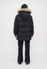 Mujer de espaldas, vestida con una chaqueta acolchada negra con capucha de pelo, y jeans oscuros de campana, de pie frente a un fondo liso.