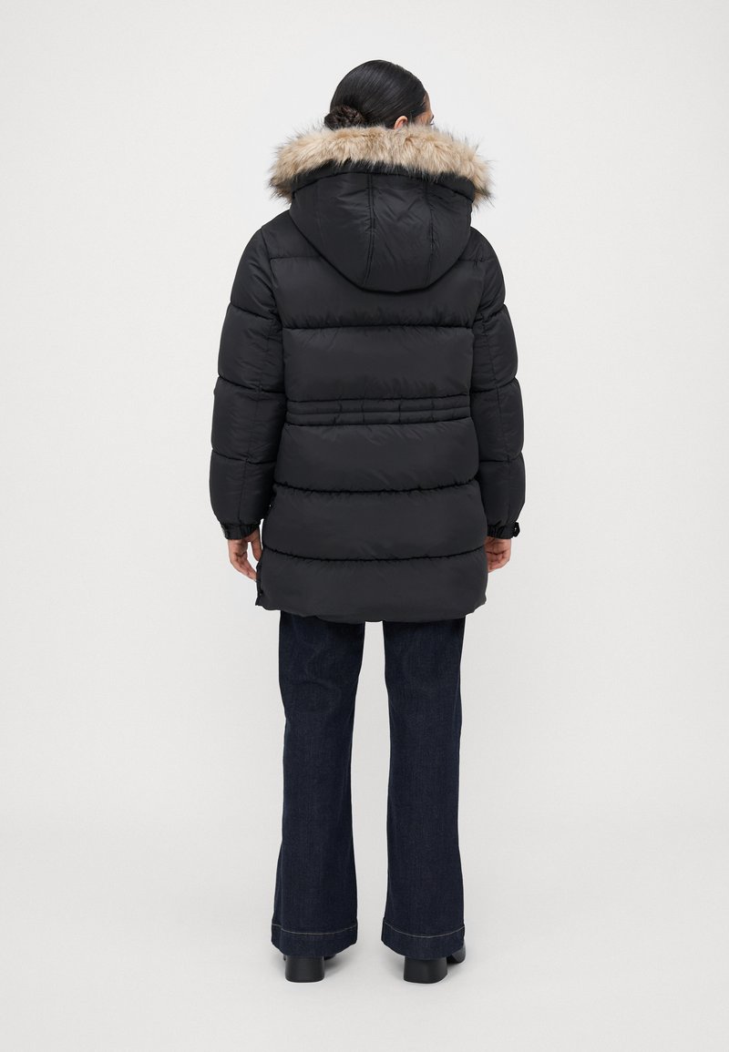 Mujer de espaldas, vestida con una chaqueta acolchada negra con capucha de pelo, y jeans oscuros de campana, de pie frente a un fondo liso.