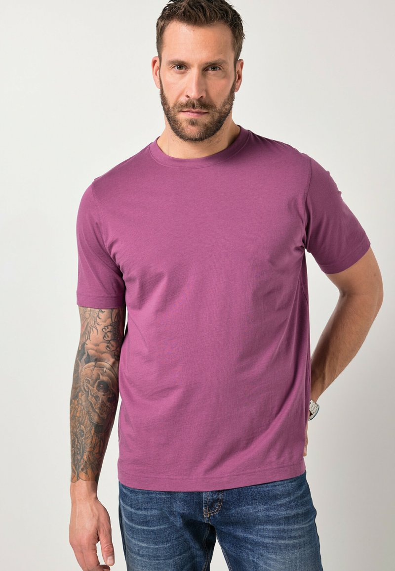 Homme avec une barbe et un bras gauche tatoué, portant un T-shirt violet et un jean bleu, debout avec une main derrière le dos.