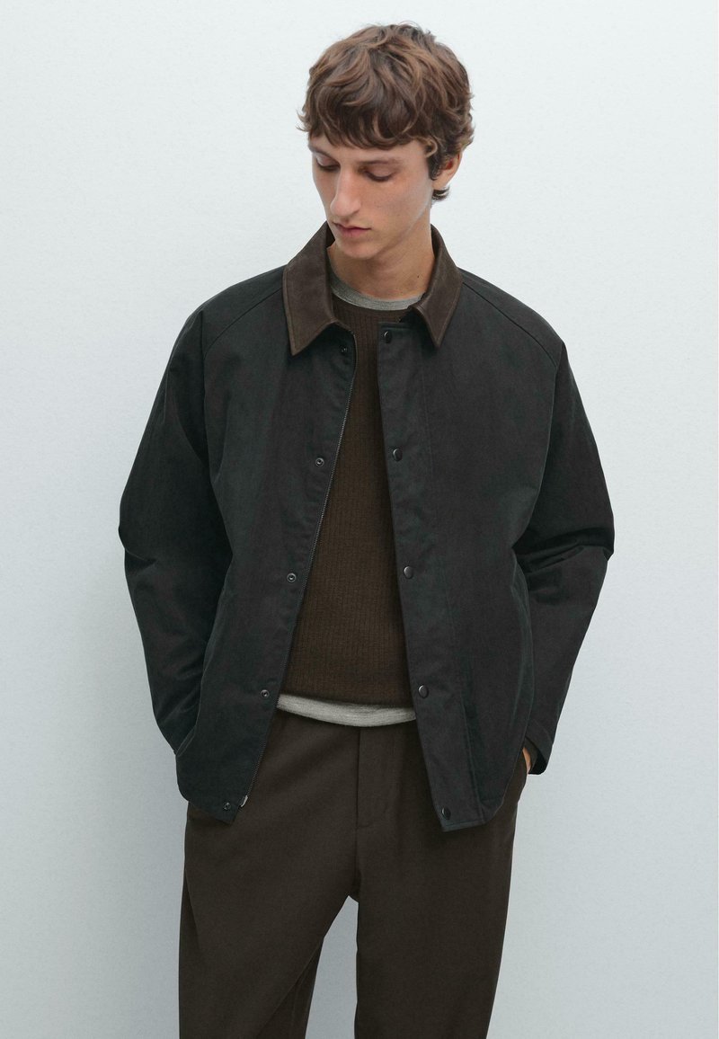 Mørk jakke med en brun krave, der har trykknapslukninger og lange ærmer. Båret over en brun sweater, mod en ensfarvet baggrund.