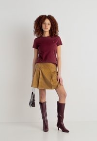 Γυναίκα με μπλουζάκι μπορντό, μίντι πλισέ φούστα καρό μουσταρδί, μπότες μέχρι το γόνατο σε τόνους μπορντό που ταιριάζουν, κρατώντας ένα μικρό πορτοφόλι με φουλάρι με σχέδιο.